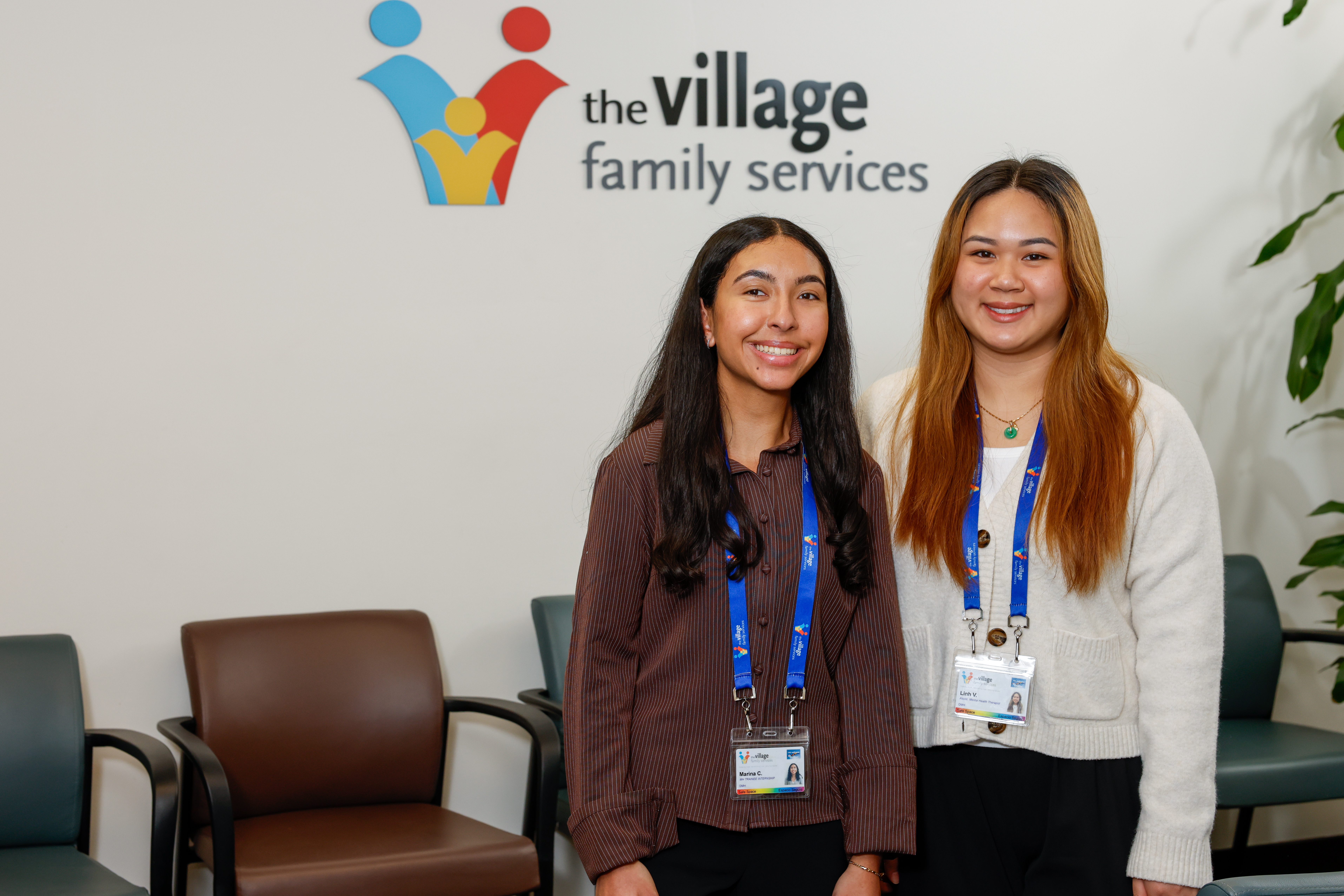 A photo of two of The Village Family Services Team members standing in our office in front of the agency logo on the wall