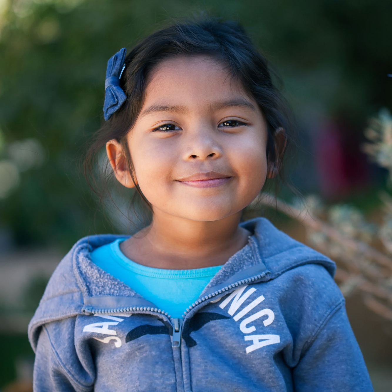 Confident little girl wearing a sweater, standing in front of trees and green plants with a sincere smile on her face.