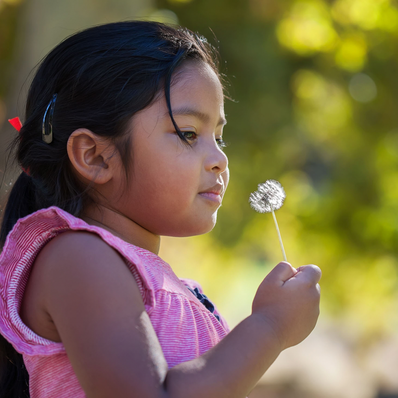 Behavioral Health Page | A thougthful little kid holding a dandelion in her hand that is close to her face and surrounded by green foliage.